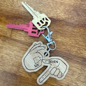 May include: A wooden keychain with a silver clasp, featuring a design of two hands forming the numbers six and seven in American Sign Language. Two keys are attached to the keychain, one gold and one pink.