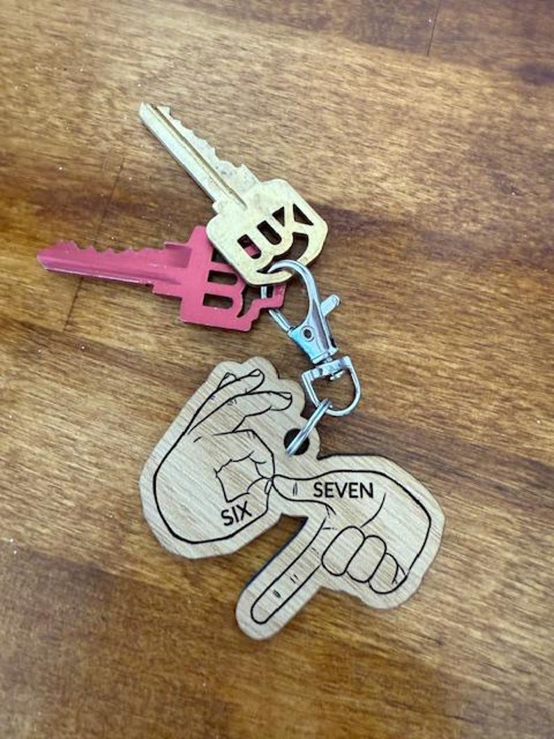 May include: A wooden keychain with a silver clasp, featuring a design of two hands forming the numbers six and seven in American Sign Language. Two keys are attached to the keychain, one gold and one pink.
