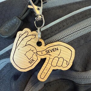 May include: Wooden keychain with a silver clasp. The keychain is shaped like two hands forming the number seven in sign language. The word "SIX" is on one hand, and the word "SEVEN" is on the other.