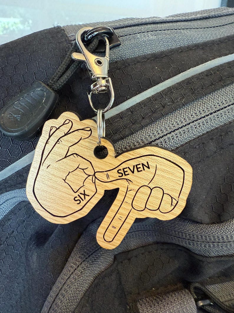 May include: Wooden keychain with a silver clasp. The keychain is shaped like two hands forming the number seven in sign language. The word "SIX" is on one hand, and the word "SEVEN" is on the other.
