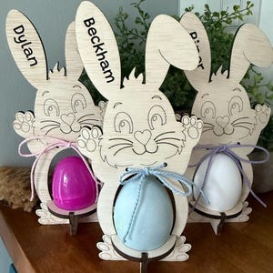 May include: Wooden Easter bunny decorations with cut-out faces and ears, holding colorful eggs. The names "Dylan", "Beckham", and "D" are written on the bunny ears. Each bunny has a different colored egg: pink, blue, and white.