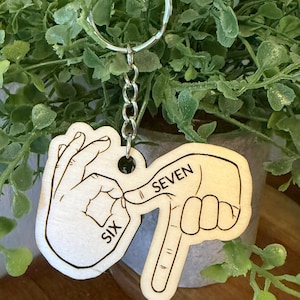 May include: A wooden keychain with a silver ring and chain. The keychain features two hand-shaped cutouts, one showing the number six in sign language and the other showing the number seven. The word "SIX" and "SEVEN" are printed on the keychain.