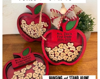 May include: Three red apple-shaped wooden ornaments with a clear window to display names. Each ornament has a hanging loop and a personalized label with the teacher's name and grade level. The text on the labels reads "Mrs. Kopas Kindergarten 2024-2025", "Mrs. Davis First Grade 2024-2025", and "Hanging and Stand Alone Versions Included".