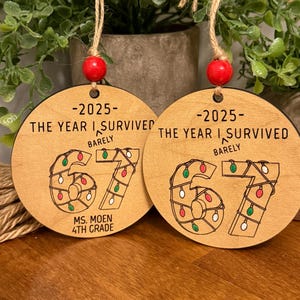 May include: Two wooden ornaments with the text "-2025- THE YEAR I SURVIVED BARELY" and "67" decorated with Christmas lights. Each ornament has a red bead and twine for hanging. The bottom of one ornament reads "MS. MOEN 4TH GRADE."