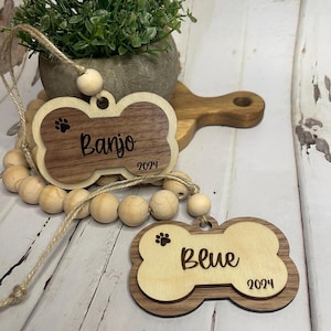 May include: Two wooden dog bone-shaped tags with engraved paw prints and names. The top tag reads "Banjo 2024" and the bottom tag reads "Blue 2024".