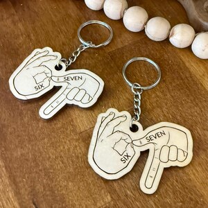 May include: Two wooden keychains with silver key rings. Each keychain features a hand gesture with the word "SIX" and "SEVEN" printed on it. The keychains are light brown and are on a wooden surface.