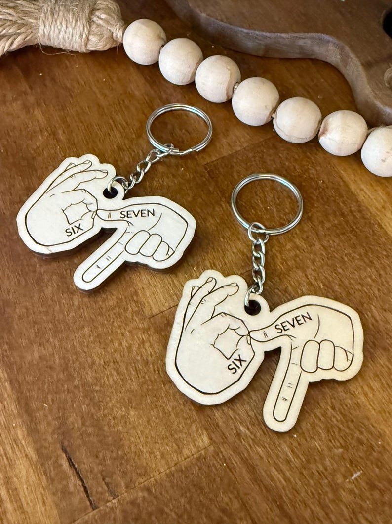 May include: Two wooden keychains with silver key rings. Each keychain features a hand gesture with the word "SIX" and "SEVEN" printed on it. The keychains are light brown and are on a wooden surface.
