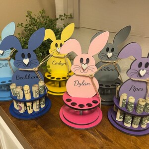 May include: Five wooden bunny money holders with personalized names, each with a different color: blue, yellow, pink, gray, and purple. Each bunny has a small heart on its nose and is holding rolled up bills.
