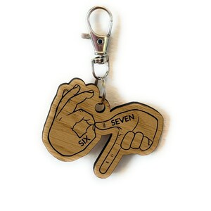 May include: A wooden keychain with a silver clasp. The keychain is shaped like two hands, one forming the number six and the other forming the number seven in American Sign Language. The words "SIX" and "SEVEN" are printed on the keychain.