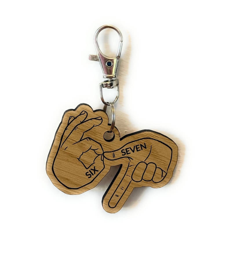 May include: A wooden keychain with a silver clasp. The keychain is shaped like two hands, one forming the number six and the other forming the number seven in American Sign Language. The words "SIX" and "SEVEN" are printed on the keychain.