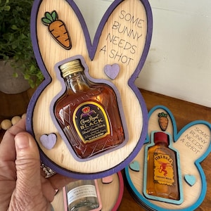 May include: Two wooden bunny-shaped coasters with a purple and blue trim. The coasters have a cutout for a mini liquor bottle. One coaster has a bottle of Crown Royal Black whiskey and the other has a bottle of Fireball Cinnamon Whiskey. The coasters have the text "Some bunny needs a shot" on them.