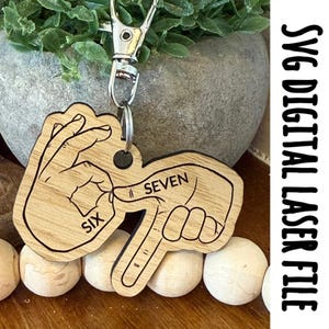 May include: Wooden keychain with a silver clasp, featuring a laser-cut design of hands spelling out the numbers six and seven in American Sign Language. The text "SVG DIGITAL LASER FILE" is printed vertically on the right side.