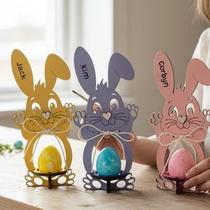May include: Three wooden Easter bunny decorations in yellow, purple, and pink, each holding a colorful egg. The bunnies have cut-out faces and paws, with names like "Jack," "Kim," and "Corbyn" on their ears. A decorative bow is tied around the egg holder.