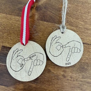 May include: Two wooden round ornaments with hand-drawn illustrations of hands using American Sign Language to spell out numbers. One ornament has "SIX SEVEN" and the other has "2025". One has a red and white ribbon, the other has a twine string.
