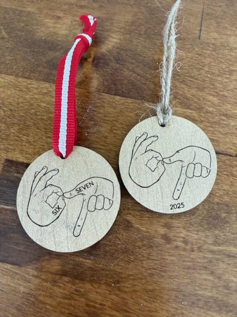 May include: Two wooden round ornaments with hand-drawn illustrations of hands using American Sign Language to spell out numbers. One ornament has "SIX SEVEN" and the other has "2025". One has a red and white ribbon, the other has a twine string.