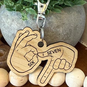 May include: Wooden keychain with a silver clasp. The keychain features a design of two hands forming the numbers six and seven in American Sign Language. The word "SIX" is printed on one hand, and "SEVEN" on the other.