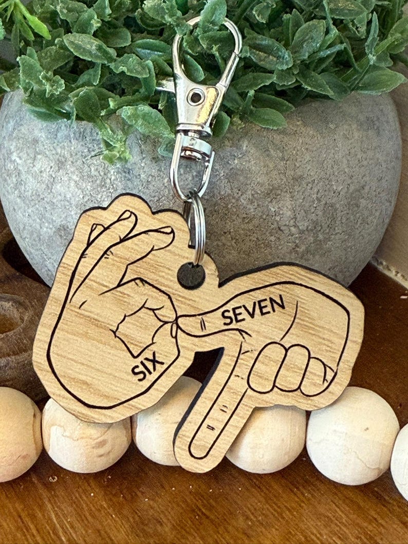 May include: Wooden keychain with a silver clasp. The keychain features a design of two hands forming the numbers six and seven in American Sign Language. The word "SIX" is printed on one hand, and "SEVEN" on the other.