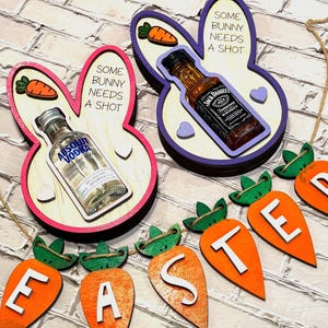 May include: Two wooden bunny-shaped ornaments with a miniature bottle of liquor inside each. The bunny on the left has a pink and white color scheme and says "Some bunny needs a shot" and the bunny on the right has a purple and white color scheme and says "Some bunny needs a shot". A wooden carrot banner with the word "EASTER" spelled out in white letters hangs below the bunnies.