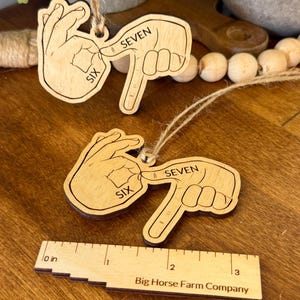 May include: Two wooden ornaments depicting hand gestures for the numbers six and seven in sign language. Each ornament is light brown with dark brown etched details and a twine loop for hanging. A wooden ruler with the text "Big Horse Farm Company" is also visible.