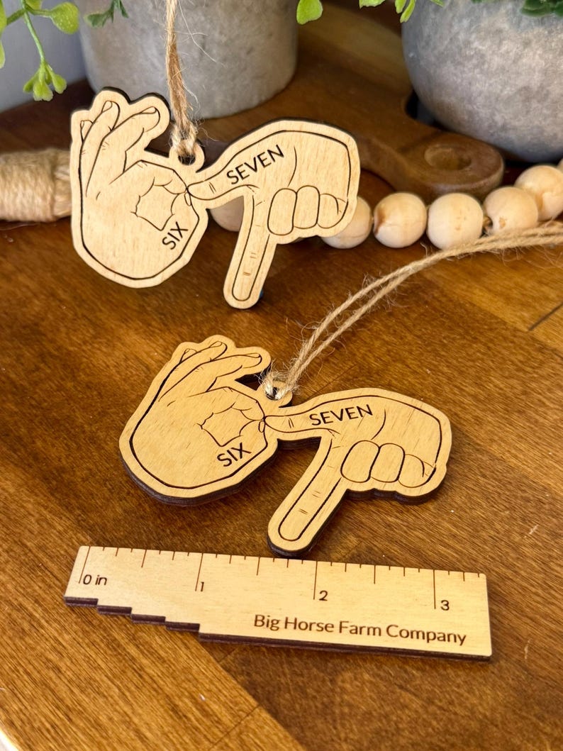 May include: Two wooden ornaments depicting hand gestures for the numbers six and seven in sign language. Each ornament is light brown with dark brown etched details and a twine loop for hanging. A wooden ruler with the text "Big Horse Farm Company" is also visible.