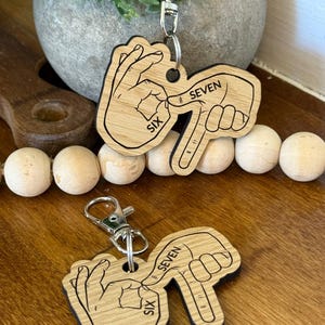 May include: Two wooden keychains with a hand gesture showing the number six and seven. The keychains are light brown with black outlines and silver key rings. The word "SIX" and "SEVEN" are printed on the keychains.