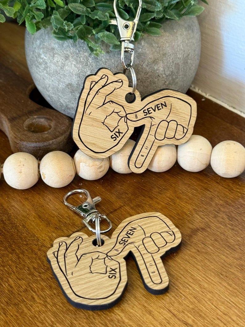 May include: Two wooden keychains with a hand gesture showing the number six and seven. The keychains are light brown with black outlines and silver key rings. The word "SIX" and "SEVEN" are printed on the keychains.