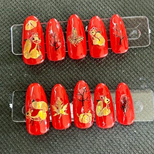 May include: A set of ten red press-on nails with autumn designs. Each nail has a unique design, including squirrels, leaves, and pumpkins. The artwork is in gold, brown, and black.