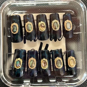 May include: A set of ten black and green press-on nails with gold accents. Each nail features a gold oval frame with a mother-of-pearl design inside.