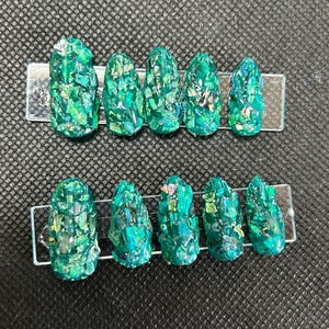 May include: Ten teal green oval-shaped false nails with a glittery finish. The nails are arranged in two rows on a clear plastic display card.