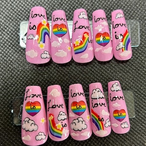 Può includere: Unghie finte rosa con disegni arcobaleno e nuvole. Ogni unghia presenta la frase "love is" in corsivo nero, insieme a un cuore arcobaleno o grafiche arcobaleno e nuvole. Le unghie sono disposte su un foglio di plastica trasparente.