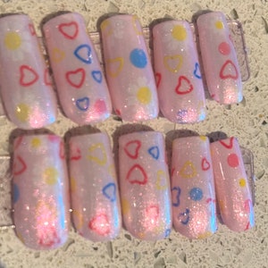 May include: Set of ten pink press-on nails with a glittery finish. The nails are decorated with small hearts, dots, and circles in various colors, including red, blue, and yellow.