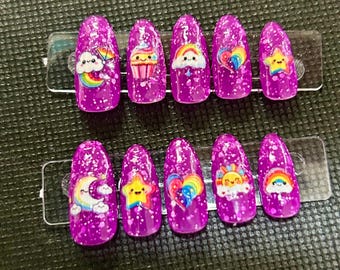 Puede incluir: Un juego de diez uñas postizas moradas con purpurina y diseños de dibujos animados coloridos. Cada uña presenta un diseño diferente, incluyendo arcoíris, nubes, corazones, estrellas y cupcakes. Las uñas son de forma ovalada y se muestran en un soporte de plástico transparente.