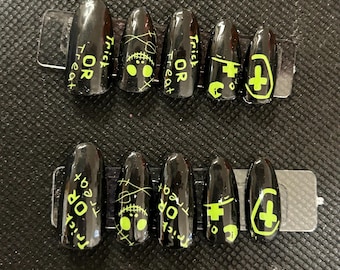 May include: Black press-on nails with neon green Halloween-themed designs. Each nail features a different design, including the words "Trick or Treat", a ghost face, and a cross symbol. The nails are oval-shaped.