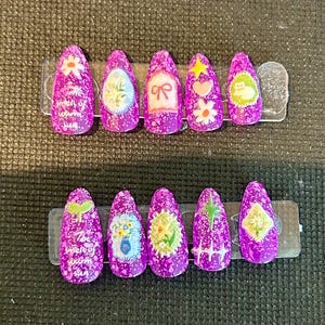 Könnte beinhalten: Ein Set aus zehn lila Press-On-Nägeln mit verschiedenen Designs. Jeder Nagel hat ein anderes Design, darunter Blumen, eine Schleife und ein Herz. Die Nägel haben ein gesprenkeltes Finish und den Text "The smell of warm sun".