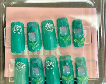 Puede incluir: Un juego de diez uñas artificiales en una caja de plástico transparente. Cada uña está pintada de color verde azulado y presenta un diseño floral blanco y un tulipán morado 3D con hojas verdes.