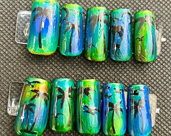 May include: Set of ten artificial nails with a tropical theme. Each nail features a palm tree design in black against a backdrop of blue, green, and yellow hues. The nails are attached to clear plastic holders.