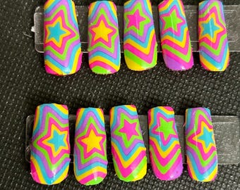 May include: Set of ten artificial nails with a vibrant, psychedelic design. Each nail features a star in the center, surrounded by concentric waves of bright colors including pink, yellow, green, blue, and purple. The nails are rectangular with rounded edges.