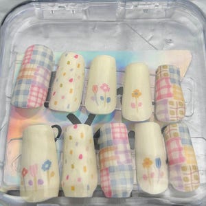 Könnte beinhalten: Ein Set aus zehn künstlichen Nägeln in einem transparenten Kunststoffbehälter. Die Nägel weisen verschiedene Designs auf, darunter pastellfarbene Karomuster, bunte Tupfen und florale Motive. Die Nägel sind in verschiedenen Formen und Größen erhältlich.