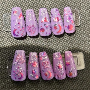 Puede incluir: Un conjunto de uñas artificiales de color lavanda con diseños temáticos del espacio. Cada uña presenta un diseño diferente, incluyendo estrellas, lunas, planetas y formas geométricas, acentuado con purpurina plateada y detalles rosas.