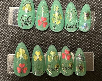 May include: Set of ten green and glitter press-on nails with shamrock and clover designs. Some nails have the words "lucky you" and "luck" written on them.