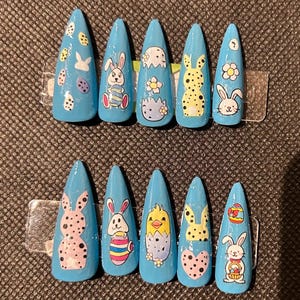 Könnte beinhalten: Satz mit zehn blauen falschen Nägeln mit Ostern-Motiven. Die Designs beinhalten Hasen, Eier, Küken und Blumen.