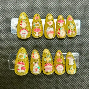 Puede incluir: Un juego de diez uñas postizas amarillas con acabado brillante. Cada uña presenta un diseño caprichoso diferente, que incluye hongos, conejitos y detalles florales. Las uñas son de forma ovalada y tienen un acabado brillante.