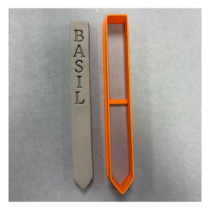 May include: A light gray, elongated, pointed garden marker with the word BASIL embossed vertically in capital letters, positioned next to a matching bright orange plastic mold or cutter with a central horizontal support bar.