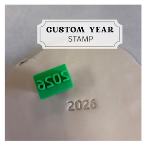 May include: A rectangular green stamp featuring the text 2026 in a mirrored orientation. The stamp is positioned next to an impression of the same text, 2026, pressed into a light-colored clay surface. The image includes the text CUSTOM YEAR STAMP.