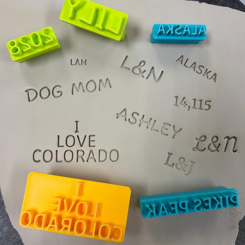 May include: A collection of plastic stamps in bright yellow, lime green, and cyan blue colours. The stamps feature various text imprints including LILY, 2028, ALASKA, DOG MOM, L&N, 14,115, ASHLEY, L&J, I LOVE COLORADO, and PIKES PEAK pressed into clay.