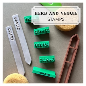 May include: A set of green rubber stamps with the text HERB AND VEGGIE STAMPS, featuring individual stamps labeled TOMATO, POTATO, GARLIC, and CELERY. Includes two light gray plant markers, a brown frame, and two round yellow sponges.