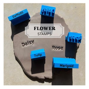 May include: A set of five bright blue plastic stamps for clay, each featuring a different flower name: Daffodil, Daisy, Tulip, Rose, and Marigold. The stamps are arranged around a central piece of brown clay, which shows the imprinted words Daisy and Rose.