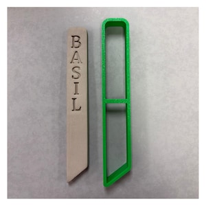 May include: A light beige rectangular plant marker with the word BASIL embossed vertically in capital letters, paired with a matching bright green plastic cookie cutter frame that mirrors the shape of the plant marker.