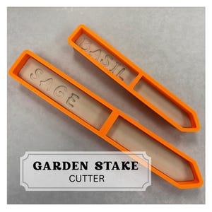May include: Two orange plastic garden stake cutters. One cutter is labeled "SAGE" and the other is labeled "BASIL". A white sign at the bottom reads "GARDEN STAKE CUTTER".