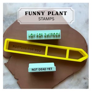 May include: A yellow plastic plant marker cutter with the text ROOTING FOR YOU, accompanied by two light green rectangular stamps. One stamp features the text ROOTING FOR YOU in reverse, and the other features the text NOT DEAD YET.